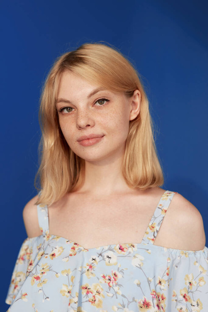 Closeup front view of an adorable blonde young woman with blonde hair, looking at camera, isolated blue background. - Zdjęcie, obraz