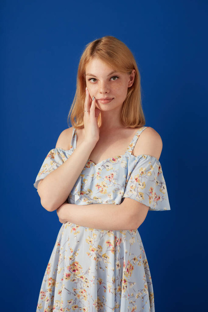 Beautiful young woman with fair hair and freckles, dressed casually, looking at camera, holding a hand near face, isolated blue background. - Φωτογραφία, εικόνα