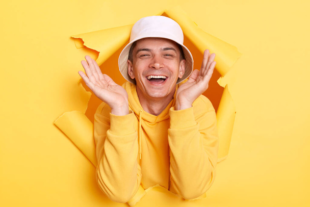 Image of pleased extremely happy man wearing hoodie and panama posing in hole in yellow paper wall, raised arms and being pleasantly surprised, looking at camera with toothy smile. - Фото, изображение