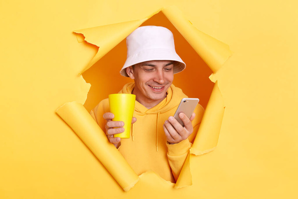 Horizontal shot of positive smiling man wearing hoodie and panama, holding cup and using cell phone, checking social networks, breaking through torn in yellow paper wall. - Valokuva, kuva