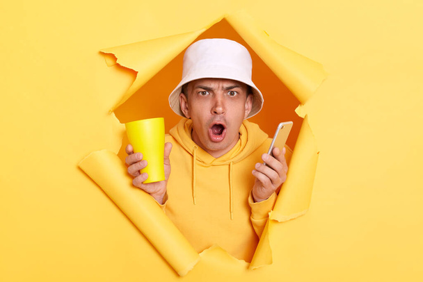 Horizontal shot of amazed surprised man wearing hoodie and panama breaking through torn in yellow paper wall, looking at camera with big eyes and open mouth, having scared expression. - Foto, imagen