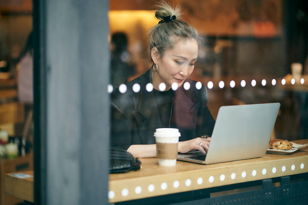 μέσω γυάλινη λήψη μιας μεσήλικας Ασιάτισσας επιχειρηματία που εργάζεται σε καφετέρια χρησιμοποιώντας φορητό υπολογιστή - Φωτογραφία, εικόνα