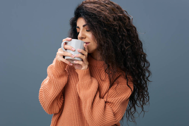 Tijd voor een kopje koffie-koffie. Genieten van gebruinde krullende Latijnse vrouw in warme trui houden kopje thee door beide handen poseren geïsoleerd over grijs blauwe achtergrond nauwe ogen. Kopieer ruimte Mockup Banner. Modeaanbod - Foto, afbeelding