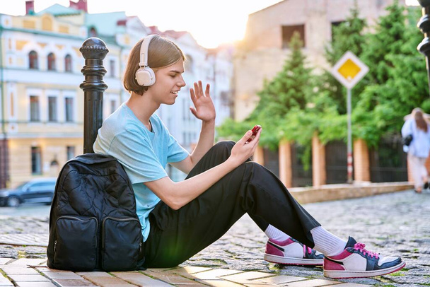 Handsome guy making video call on smartphone, outdoor on city street. Young male in headphones with backpack sitting on sidewalk talking online, video chat conference, leisure communication technology - Fotoğraf, Görsel