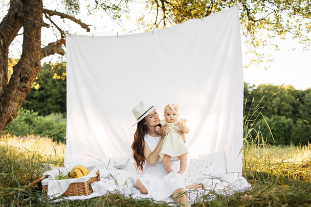 Positive caucasian mother with dark hair and white hat sitting with her little daughter on white blanket outdood. Family of two enjoying summer camping on fresh air. - Foto, afbeelding