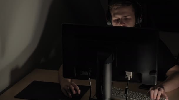 Man playing computer games in a dark room. Using keyboard and mouse. Wearing headphones. Caucasian ethnicity. Display. White and black wall. Beige table. Modern technology. - Footage, Video