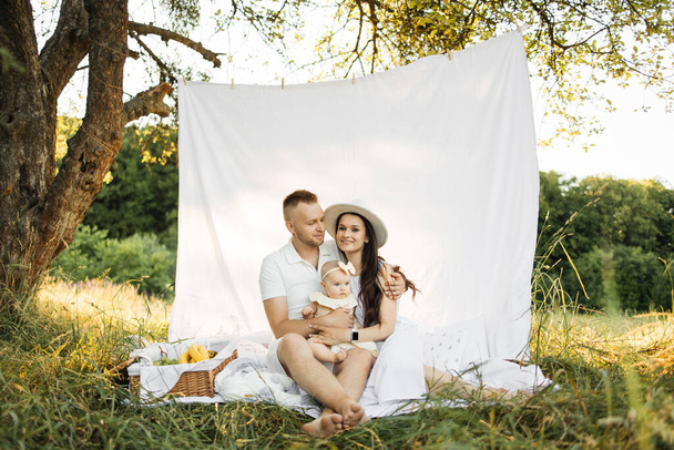 Beautiful caucasian man and woman sitting in embrace with their cute little daughter on white blanket with curtain. Family time on fresh air. - Фото, изображение