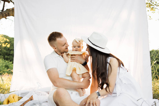 Portrait of young parents in white wear family look playing with six months old daughter on nature holding her in hands. Concept of family time and outdoors activity. Copy space. - Foto, afbeelding