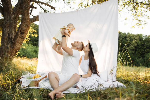 Mooie en gelukkige blanke ouders die spelen met hun zes maanden oude dochter die op een witte deken zit. Positieve familie van drie genieten van vrije tijd op frisse lucht. - Foto, afbeelding