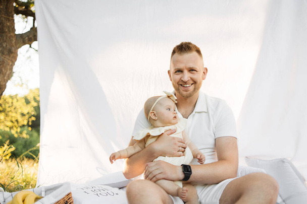 Blond haired cheerful happy man relaxing on white blanket with curtain in park with little baby girl while looking at camera. Family summer concept. - Фото, изображение