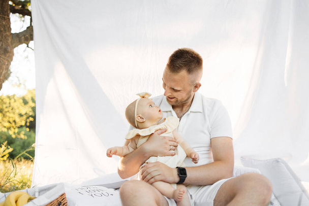 Blond haired cheerful happy man relaxing on white blanket with curtain in park with little baby girl while looking at camera. Family summer concept. - Photo, Image