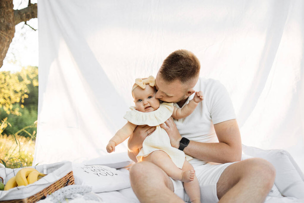 Happy caucasian father kissing and hugging his lovely little daughter while sitting near white tent on fresh air. Concept of parenting, fun and leisure time. - Foto, afbeelding