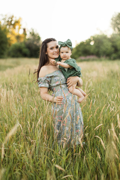 Cheerful young woman in green summer dress lifting up six month old baby girl while playing walking on field. Mother with daughter enjoying time spending together on fresh air. - Foto, Imagem
