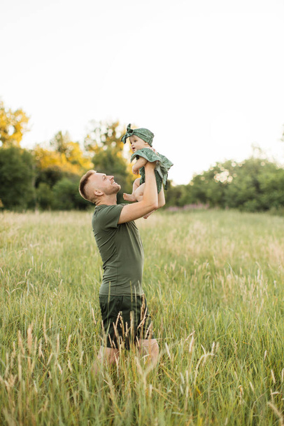 Attractive young father with green t-shirt and shorts lifting up his cute little daughter on hands playing together walking on meadows. Family time outdoors. - 写真・画像