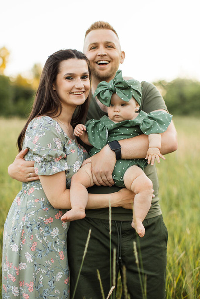 Joyful young parents playing with cute daughter outdoors while standing on background of green meadow. Man and woman holding baby girl on hands. Copy space. - Valokuva, kuva