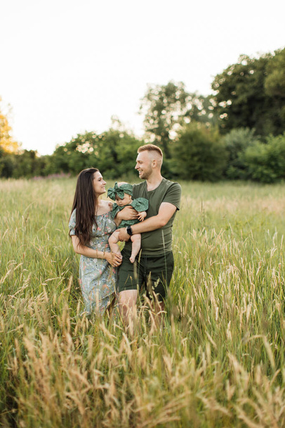 Beautiful and happy caucasian parents playing with their six months old daughter walking on green field. Positive family of three enjoying leisure time on fresh air. - Zdjęcie, obraz