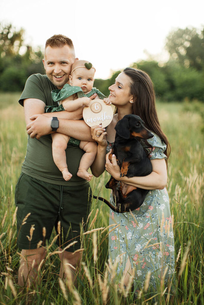 Portrait of happy young parents embracing little cute daughter and their dog celebrate six months since the birth of a child while standing on the field on fresh air. - Foto, immagini