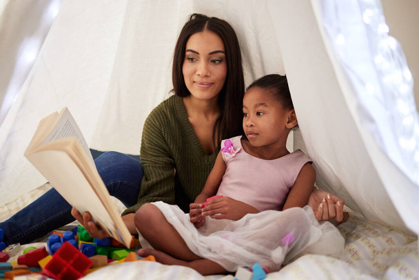 een moeder leest een boek voor aan haar dochtertje onder een deken fort thuis. - Foto, afbeelding