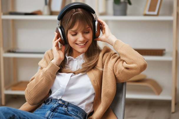 Cheerful young blonde student lady freelancer in warm sweater in headphones enjoy good playlist listen fav songs sitting in armchair at modern home interior. Music time Relaxing Concept. Copy space - Photo, Image