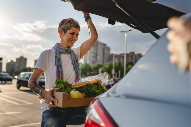 One woman mature caucasian female standing by the back trunk of her car on the parking lot of the supermarket shopping mall or grocery store with vegetables food in box putting them in the vehicle - Photo, Image