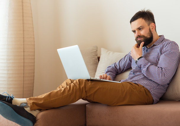 Молодий бородатий чоловік працює на ноутбуці вдома
 - Фото, зображення