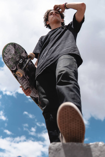 It's a low angle view of a skater on a ramp. The boy wears a black t-shirt, jeans and tennis shoes. He has an urban style. He holds his skateboard in his right arm. - Foto, Imagen