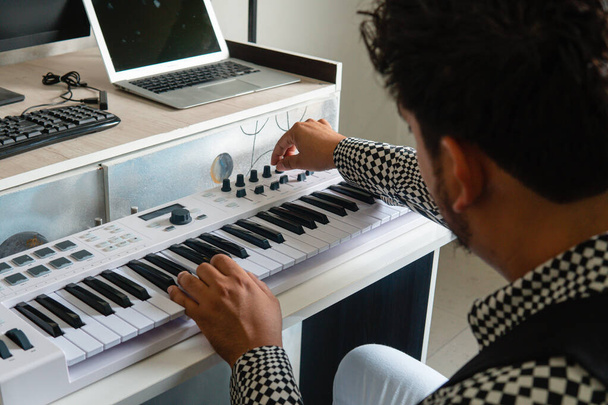 Музикант працює з клавіатурою та ноутбуком збоку. Він у чорно-білій сорочці. Його музична клавіатура біла. Це близький постріл музиканта
.  - Фото, зображення