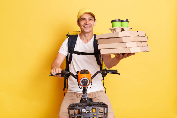 Delighted Caucasian courier man on cycle holding carton pizza boxes and disposable coffee cups, fast food delivering from restaurant, wearing white t shirt and cap, isolated over yellow background. - Foto, Imagem