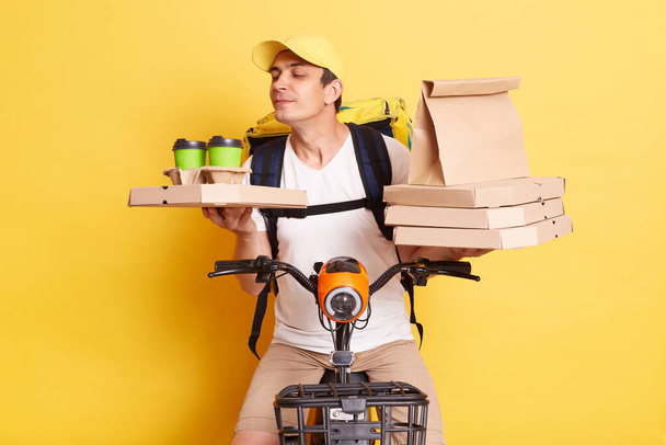 Satisfied deliveryman on bicycle holding paper parcel, pizza boxes and disposable coffee cups, smelling hot beverage, enjoying aroma while delivering food, isolated over yellow background. - Fotoğraf, Görsel