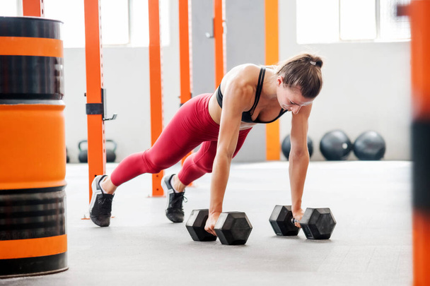 Сильна спортсменка в активному одязі з хвостом робить дошку з гантелями під час функціональних тренувань у просторому легкому тренажерному залі
 - Фото, зображення