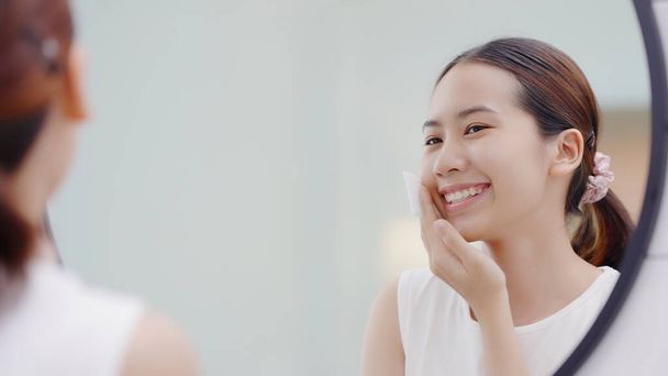 Happy Asian girl looking in to mirror, holding cotton pad, applying facial wipe on her face, removing makeup. - 写真・画像