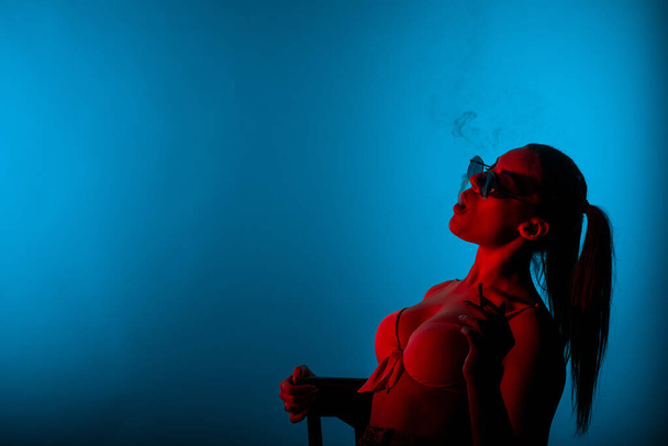 Schöne Mädchen raucht Zigarette im Studio, während sie auf einem blauen Hintergrund sitzt - Foto, Bild