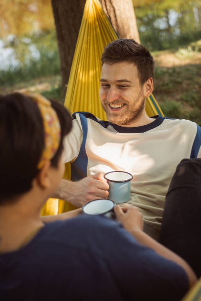 Zbliżenie ładnej pary pijącej kawę i relaksującej się na żółtym hamaku w parku - Zdjęcie, obraz