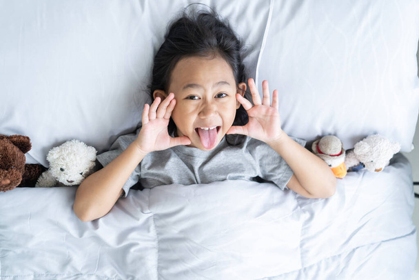 Top view cute asian child little girl wake up and lies in the bed with a toy teddy bear - 写真・画像