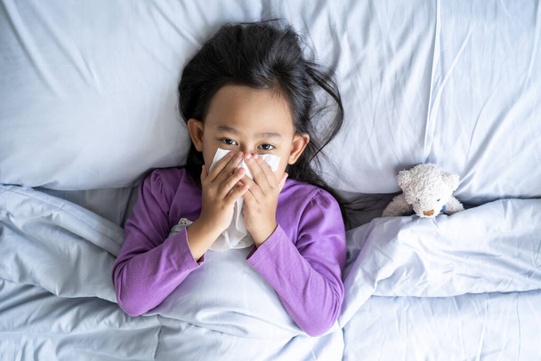 Top view Asian child girl sick with sneezing on the nose and cold cough on tissue paper because weak, Sick child with fever and illness in bed. - Zdjęcie, obraz