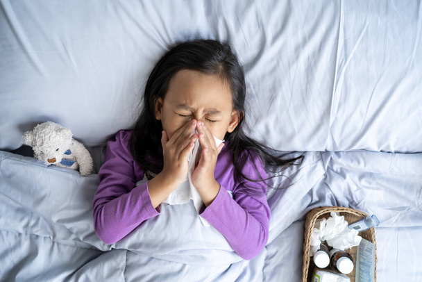 Top view Asian child girl sick with sneezing on the nose and cold cough on tissue paper because weak, Sick child with fever and illness in bed. - Photo, Image