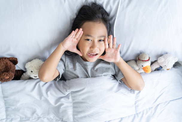 Top view χαριτωμένο ασιατικό κοριτσάκι ξυπνήσει και βρίσκεται στο κρεβάτι με ένα παιχνίδι αρκουδάκι - Φωτογραφία, εικόνα
