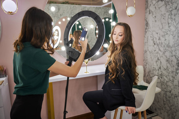 Maskenbildner fotografiert Kunden im Schönheitssalon. Junge Frau mit langen Haaren sitzt auf Stuhl und schaut Schönheitsspezialistin an, während Arbeiterin mit Smartphone fotografiert. - Foto, Bild