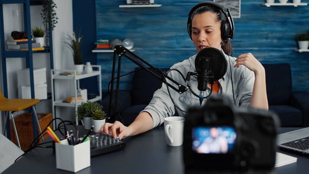 Attractive social media influencer speaking to audience at microphone while tweaking sound using professional mixer. Creative digital content creator recording podcast at home studio. - Foto, afbeelding