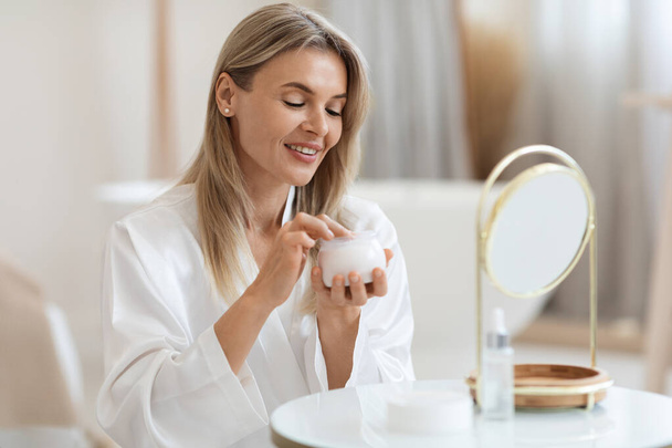 Happy middle aged pretty blonde woman in bathrobe sitting in front of mirror, holding jar with beauty product, moisturizing face cream, enjoying her morning face care routine, copy space - Photo, image