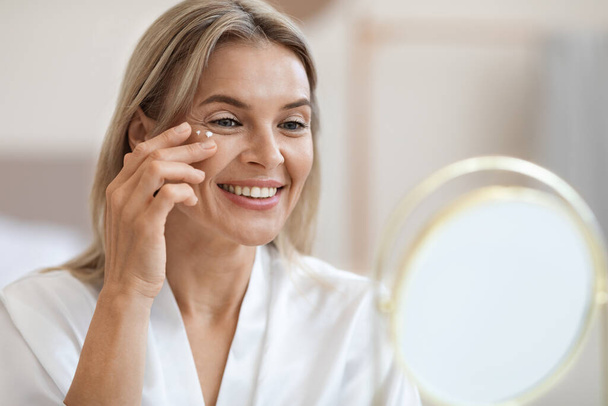 Closeup of cheerful beautiful blonde middle aged lady in silky bathrobe applying moisturizing eye cream, looking at mirror and smiling, enjoying face care routine, copy space - 写真・画像