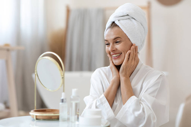 Happy beautiful middle aged woman in white bathrobe and with turban on her head looking at mirror, touching her face skin and smiling, enjoying the way she looks, home interior, copy space - Foto, Bild