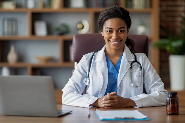 Весела лікарка молода афроамериканка в білому халаті зі стетоскопом, що працює в клініці, сидить на робочому столі, використовує ноутбук, консультує своїх пацієнтів онлайн, посміхається на камеру, копіює простір
 - Фото, зображення
