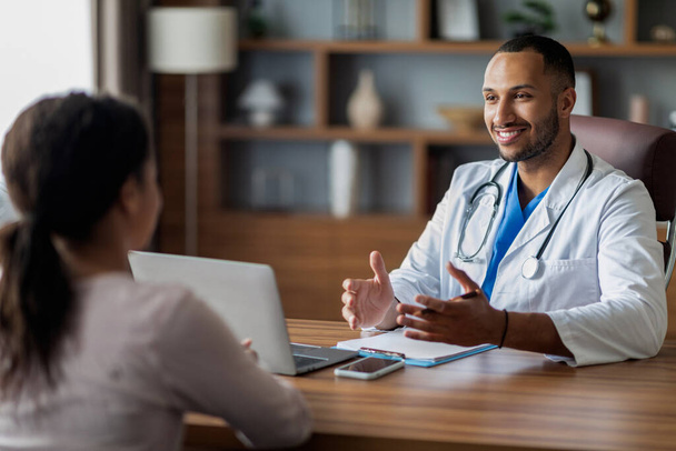 Pozytywny przystojny arabski mężczyzna lekarz konsultujący nierozpoznawalny afrykański amerykański pacjent kobieta, Doc daje rekomendacje, uśmiech i gestykulacji, siedzi przy biurku z laptopem w klinice - Zdjęcie, obraz
