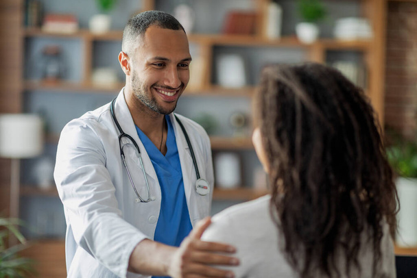 Gezondheidszorg concept. Vriendelijk aantrekkelijke Midden-Oosterse jonge man arts begroeten zijn vrouwelijke patiënt Afro-Amerikaanse vrouw, aanraken van haar schouder en glimlachen, kliniek interieur, close-up schot - Foto, afbeelding