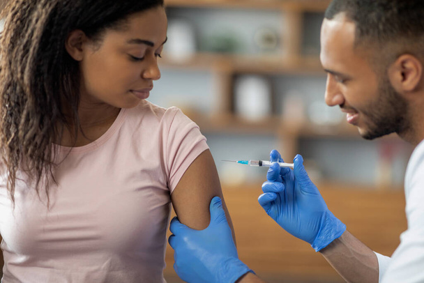 Mladá afro-americká žena, očkovaná proti koronaviru, dívá se na mužského lékaře pohledného muže ze Středního východu, jak si píchá injekci do ramene, interiér kliniky, detailní záběr, kopírovací prostor - Fotografie, Obrázek