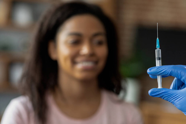 Selective focus on syringe in doctor hand in blue medical glove over blurred smiling young black woman patient, african american lady getting vaccination against COVID or flu at clinic - Photo, image