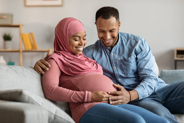 Портрет щасливої чорношкірої ісламської пари, що очікує дитину, відпочиває разом на дивані, усміхаючись молода вагітна мусульманська пара зв'язування вдома, люблячий чоловік ніжно торкається дружин Живіт, Крупним планом
 - Фото, зображення
