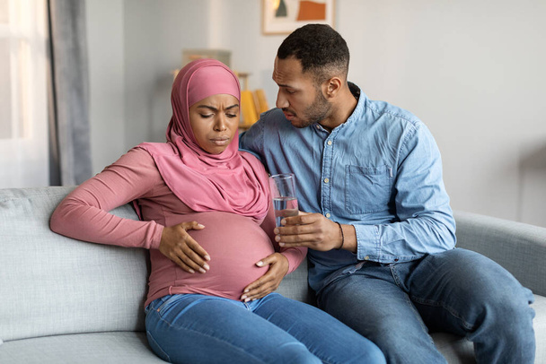 Pečující manžel podporovat jeho černé těhotné muslimské manželky, dávat jí sklenici vody, zatímco ona se cítí špatně doma, islámská žena sedí na gauči v obývacím pokoji, trpí prenatální kontrakce - Fotografie, Obrázek