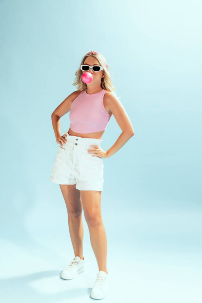 Portrait of stylish young woman in trendy sunglasses posing with bubble gum isolated on light blue studio background. Concept of beauty, emotions, facial expression, lifestyle, fashion, youth culture - 写真・画像